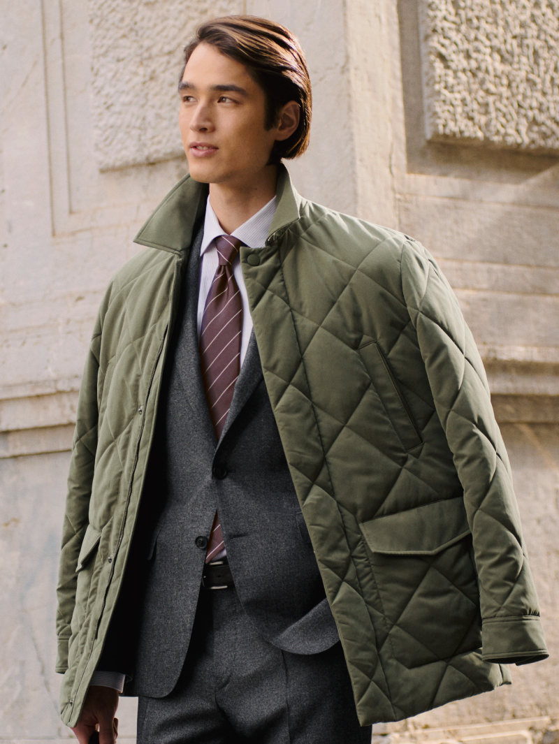 Canali model wearing a grey suit with shirt and tie under an olive green quilted jacket outdoors.