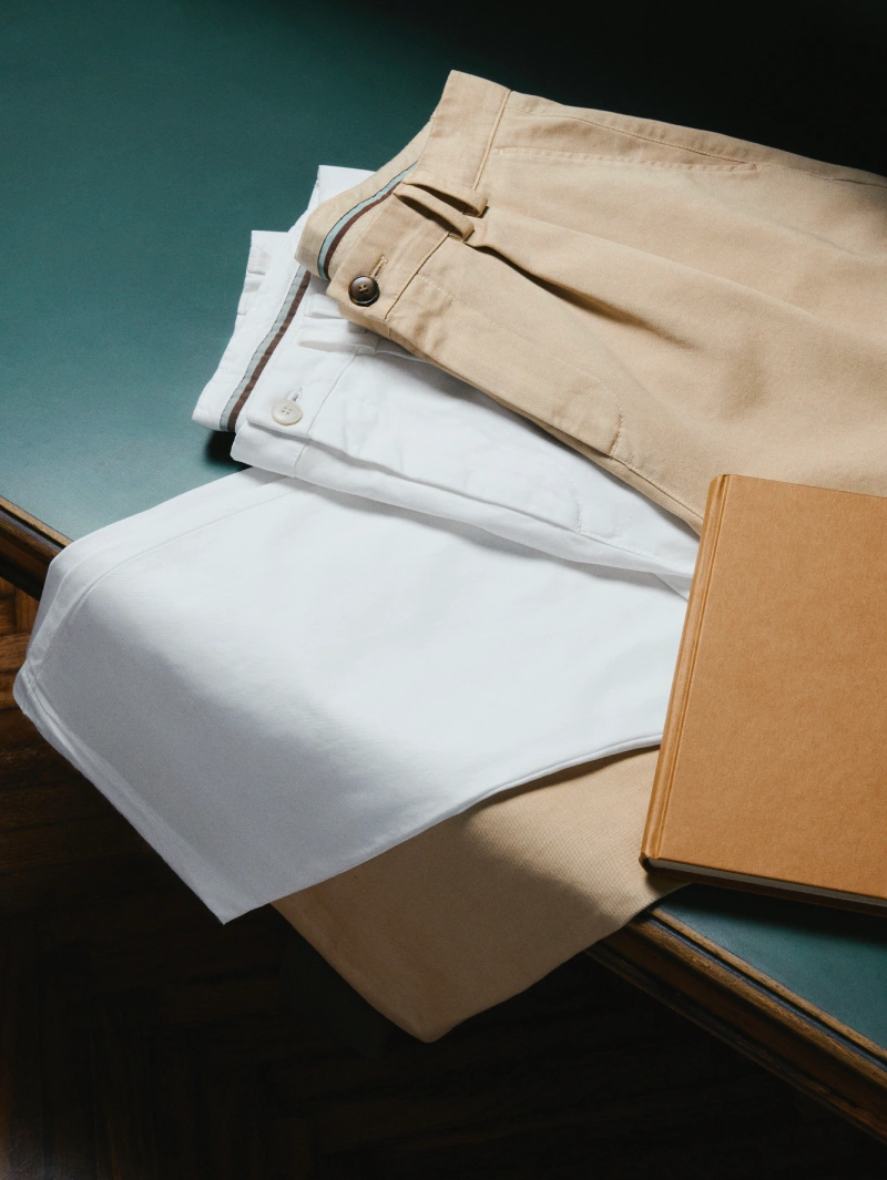 Fall/Winter 2026 beige casual trousers paired with a white shirt and knitwear, neatly folded on a wooden desk with a hardcover book, emphasizing refined everyday style.