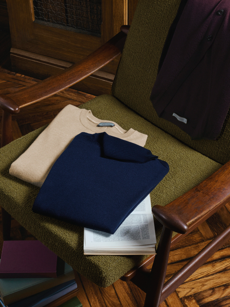 Canali menswear knitwear: a beige crew-neck sweater and a blue knitted polo folded on a green armchair beside an open book, in a warm and refined setting.